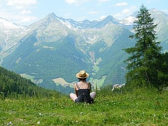 Junge Frau sitzt in einer Wiese vor einer bergigen Landschaft