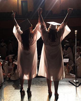 Absolventinnen stehen mit nach oben gestreckten Händen auf einer Bühne