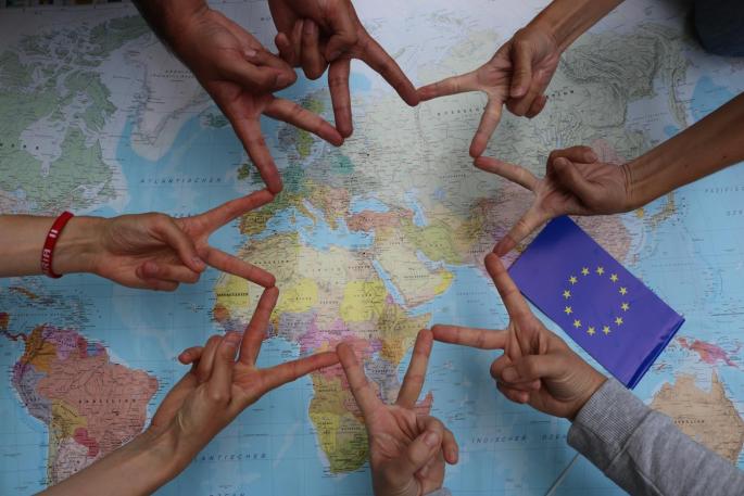 Hände die zusammen einen Stern bilden. Im Hintergrund sieht man eine Landkarte und die EU-Flagge